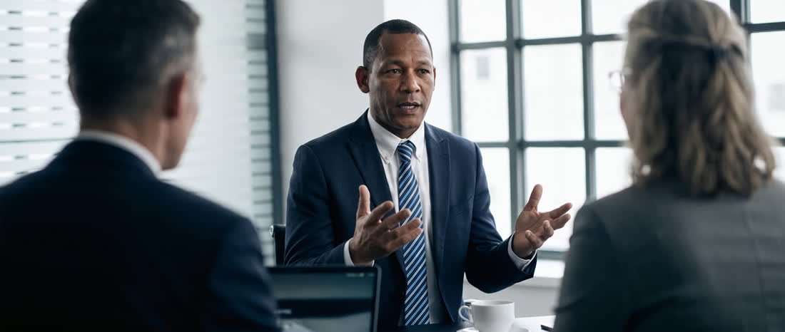 The principal mid-explanation across a table in a bright daylight office.